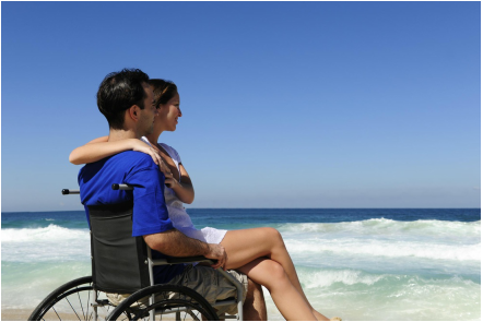 wheelchair man with woman in his lap on the beach looking at waves